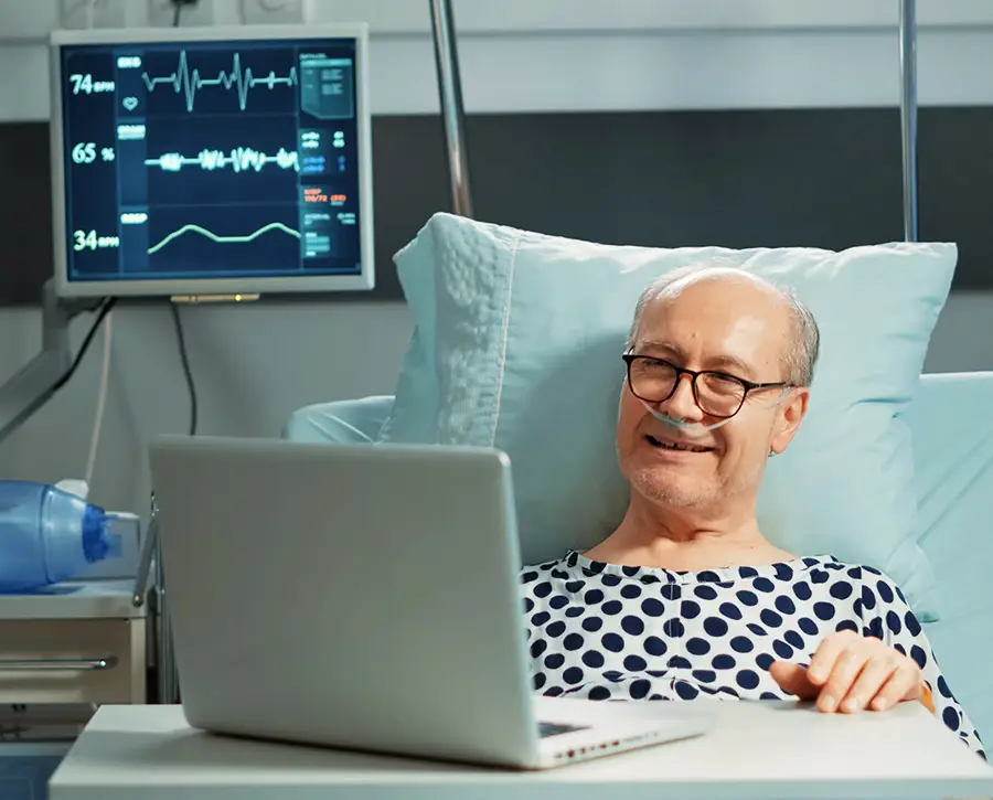 Senior in hospital bed watching funeral live stream on a laptop.
